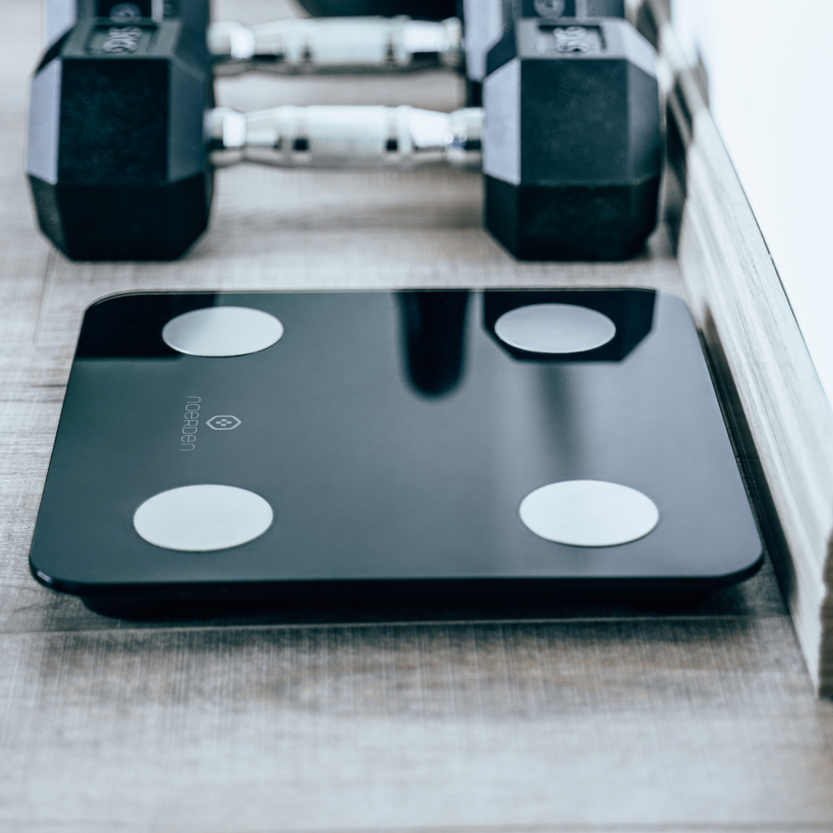 Black MINIMI smart body scale on wooden floor with weights on the floor in the background